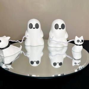 Pode incluir: Tr&ecirc;s estatuetas de cer&acirc;mica branca sobre uma superf&iacute;cie espelhada. Um fantasma com olhos pretos est&aacute; segurando um cord&atilde;o branco preso a uma estatueta de cachorro branca. Outro fantasma com olhos pretos est&aacute; segurando um cord&atilde;o branco preso a uma estatueta de gato branca.