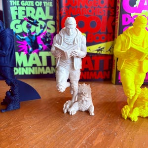 May include: A collection of 3D-printed figurines, including a white figure reading a book, a blue figure, and a yellow figure. The figurines are in front of book covers with text including "The Gate of the Feral Gods" and "The Dungeon Anarchist's Cookbook".