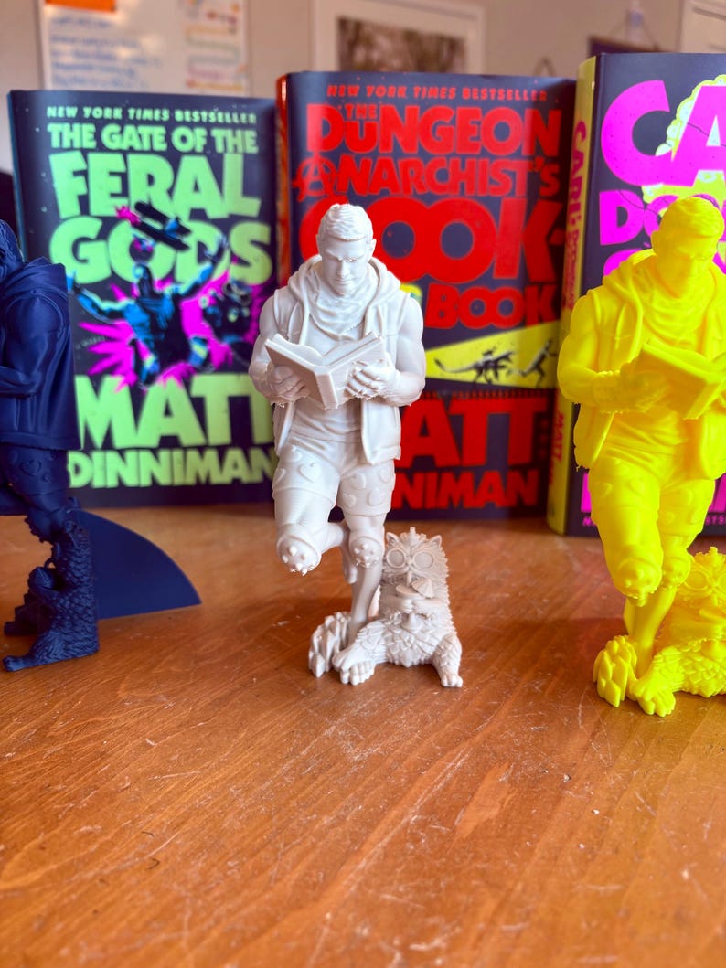 May include: A collection of 3D-printed figurines, including a white figure reading a book, a blue figure, and a yellow figure. The figurines are in front of book covers with text including "The Gate of the Feral Gods" and "The Dungeon Anarchist's Cookbook".