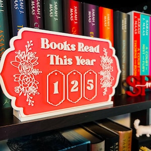 May include: A red and white decorative sign that reads "Books Read This Year" with the numbers "125" displayed. The sign is decorated with floral designs and is displayed on a bookshelf with books.