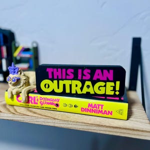 May include: A whimsical figurine of a lion wearing glasses and a unicorn horn rests on a yellow book. A black sign with the words "THIS IS AN OUTRAGE!" in pink and yellow sits atop the book. The book is titled "Dungeon Crawler Carl Book Two".