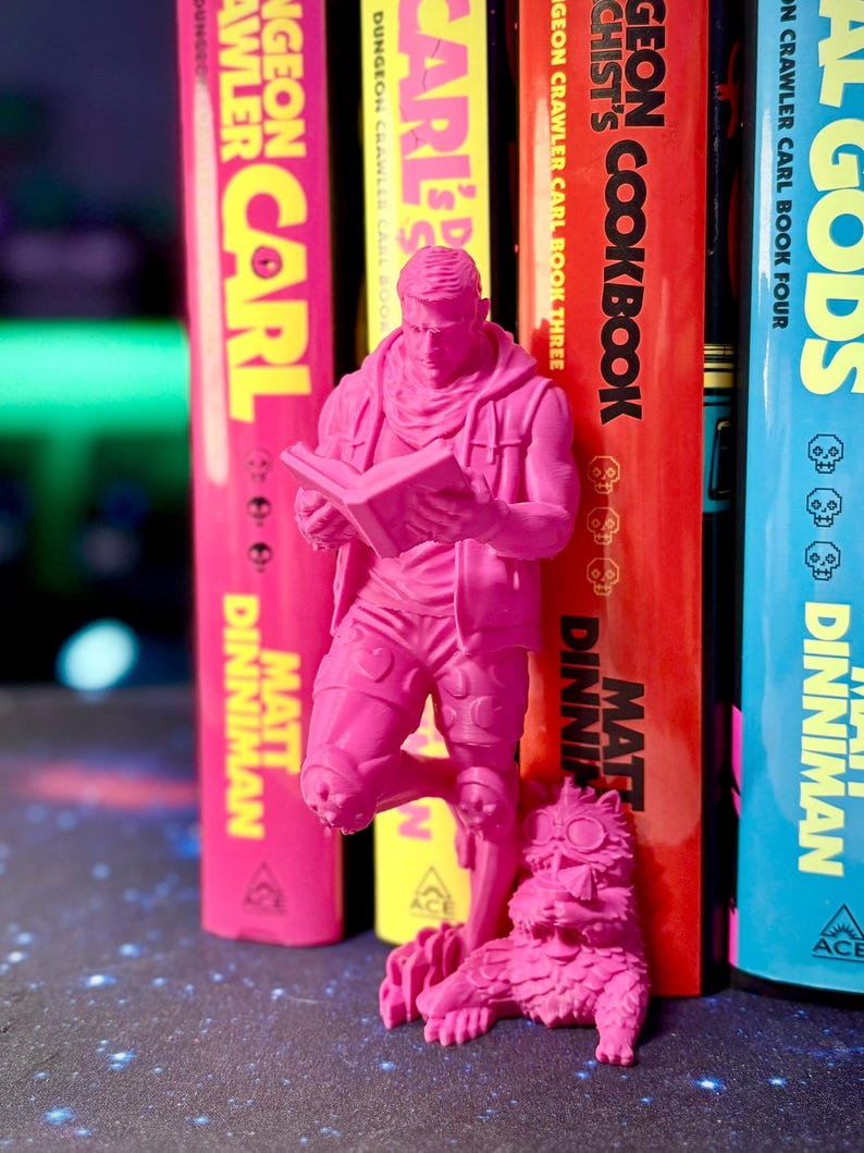 May include: A vibrant pink 3D-printed figurine of a person reading a book, positioned in front of a row of colorful books. The books have titles like "Dungeon Crawler Carl" and "Algods". A pink cat figurine is at the base.