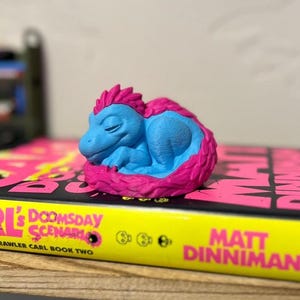 Può includere: Una piccola statuina di dinosauro addormentato, blu e rosa, è appoggiata su un libro. La statuina è blu chiaro con una cresta e un collare rosa. Il libro ha il dorso giallo con il titolo "CARL's DOOMSDAY SCENARIO".