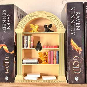 May include: A small, cream-colored bookshelf with an arched top, filled with books and decorative figurines. The bookshelf is flanked by large books with the author's name "Raven Kennedy" and book titles. The figurines include dragons and a cloaked figure.