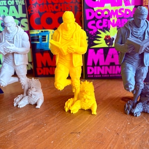May include: Three figurines of a person reading a book, each with a creature at their feet. The central figure is bright yellow, flanked by white and gray figures. Behind them are book covers with bold text in red, pink, and black.