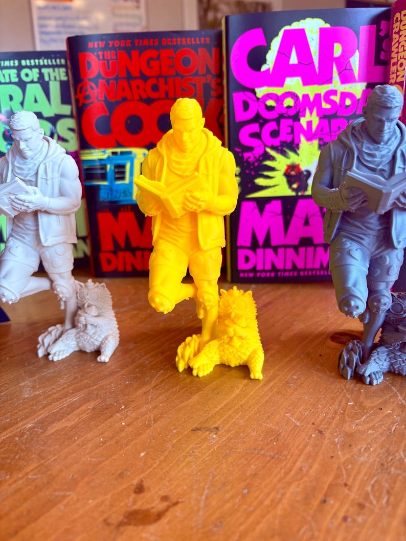 May include: Three figurines of a person reading a book, each with a creature at their feet. The central figure is bright yellow, flanked by white and gray figures. Behind them are book covers with bold text in red, pink, and black.
