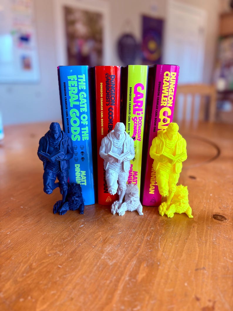 May include: Four books from the "Dungeon Crawler Carl" series stand upright, each with a different colored spine. Miniature figurines of a character and a dog are in front of each book. The books are titled "The Gate of the Feral Gods", "Anarchist's Cookbook", "Doomsday Scenario", and "Dungeon Crawler Carl".