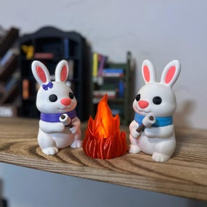 May include: Two white bunny figurines with pink ears and black eyes, wearing blue and purple scarves, sit on a wooden surface. A small, orange campfire is between them. The bunnies hold small sticks, suggesting they are roasting marshmallows.
