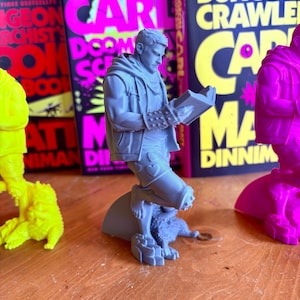 May include: A gray figurine of a man reading a book, with a dog at his feet. The figurine is in front of several books with the title "Dungeon Crawler Carl" and the author's name. The books have a black background with pink and yellow text.