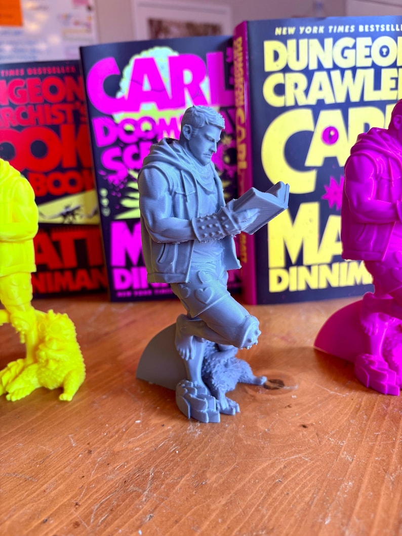 May include: A gray figurine of a man reading a book, with a dog at his feet. The figurine is in front of several books with the title "Dungeon Crawler Carl" and the author's name. The books have a black background with pink and yellow text.