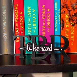 May include: A black, 3D-printed TBR (to be read) book stand. The letters are large and bold, with the words "to be read" in a cursive font. The stand is placed in front of a row of colorful books.