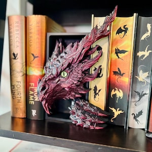 May include: A detailed, burgundy dragon head bookend with green eyes rests on a shelf next to fantasy books. The bookend is intricately sculpted, showcasing scales and sharp features. The books have titles like "Fourth Wing" and "Iron Flame".