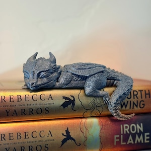 Peut inclure: Une figurine de dragon grise imprimée en 3D, posée sur une pile de livres. Le dragon a des cornes et est couché. Les titres des livres incluent "REBECCA YARROS" et "IRON FLAME".