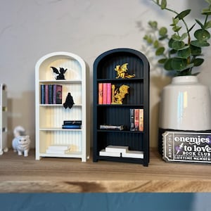 Op de afbeelding: Miniatuur witte en zwarte boekenplanken met boeken en decoratieve beeldjes. Er zijn ook een wit kattenbeeldje en een kleine rolplank. Op de plank staat een kaartje met de tekst "enemies to lovers book club".