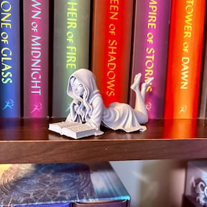 Op de afbeelding: Een lichtgrijze beeldje van een persoon die een boek leest, liggend op een plank voor een rij kleurrijke boeken. De boeken hebben titels als "Throne of Glass" en "Queen of Shadows". Het beeldje is gedetailleerd en lijkt van hars te zijn.