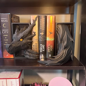 Puede incluir: Un juego de sujetalibros de dragón gris oscuro con un ojo naranja, sosteniendo libros en una estantería de madera. Los libros incluyen títulos como "Crescent City" y "Fourth Wing".