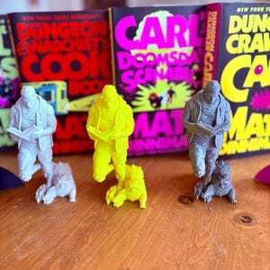 May include: Three figurines of a person reading, each with a small animal companion, are displayed in front of book covers with bold text. The figurines are white, yellow, and gray. The book covers have titles like "Dungeon Anarchist's Cook Book."