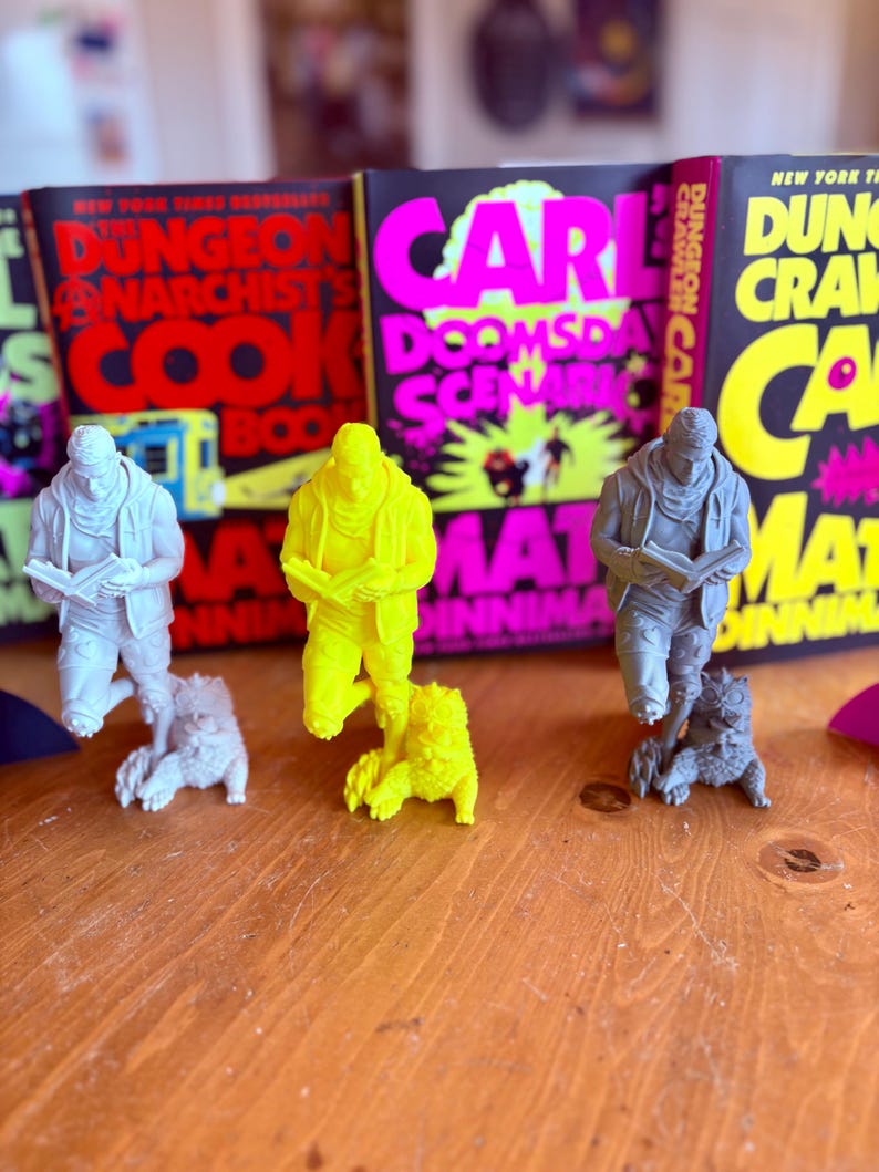 May include: Three figurines of a person reading, each with a small animal companion, are displayed in front of book covers with bold text. The figurines are white, yellow, and gray. The book covers have titles like "Dungeon Anarchist's Cook Book."