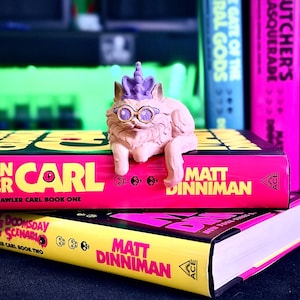K&ouml;nnte beinhalten: Eine skurrile Katzenfigur mit Brille und lila Krone sitzt auf zwei B&uuml;chern. Die B&uuml;cher tragen die Titel "Dungeon Crawler Carl" und "Doomsday Scenario" von Matt Dinniman, mit pinkfarbenen und gelben Einb&auml;nden.