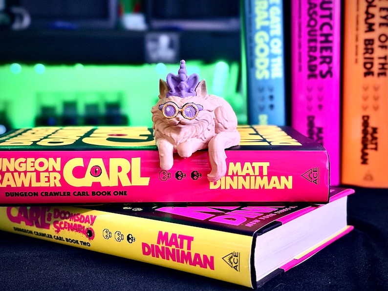K&ouml;nnte beinhalten: Eine skurrile Katzenfigur mit Brille und lila Krone sitzt auf zwei B&uuml;chern. Die B&uuml;cher tragen die Titel "Dungeon Crawler Carl" und "Doomsday Scenario" von Matt Dinniman, mit pinkfarbenen und gelben Einb&auml;nden.