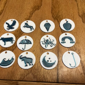 Könnte beinhalten: Zwölf weiße runde Ornamente mit schwarzen und blauen Bildern darauf. Die Bilder zeigen einen Vogel, ein Einhorn, einen Eisbecher, eine Eichel, eine Kuh, einen Regenschirm, einen Oktopus, einen Regenbogen, eine Welle, einen Elefanten, einen Halbmond und einen Angelhaken.