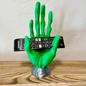 May include: A green hand-shaped remote control holder with a silver base. The holder is designed to cradle a black Samsung remote control. The fingers are outstretched, and the overall design is a unique decorative item.