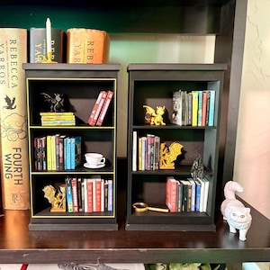 Puede incluir: Dos pequeñas estanterías negras con ribetes dorados, llenas de libros diminutos, figuritas y una taza de té. Una estantería tiene una figurita de gato blanco. Los libros son de varios colores y las figuritas incluyen dragones.
