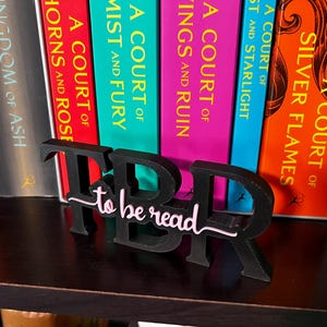 May include: A black "TBR to be read" book stand sits in front of a row of colorful books. The stand has a cursive "to be read" in pink and white. The books have titles like "A Court of Silver Flames" and "Kingdom of Ash".