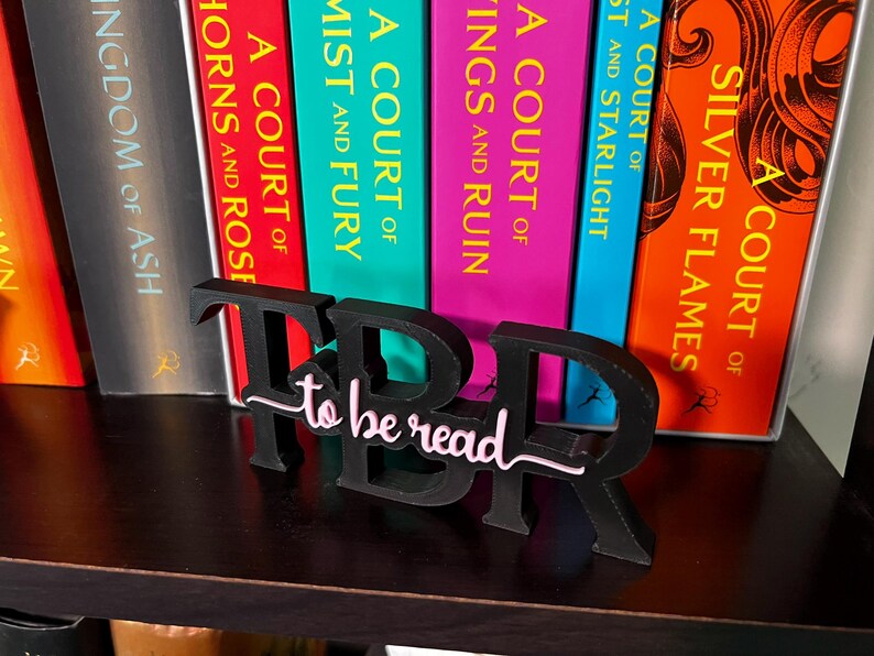 May include: A black "TBR to be read" book stand sits in front of a row of colorful books. The stand has a cursive "to be read" in pink and white. The books have titles like "A Court of Silver Flames" and "Kingdom of Ash".