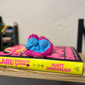 May include: A blue and pink dinosaur figurine curled up on a book. The book has a yellow spine with the text "CARL'S DOOMSDAY SCENARIO" and "MATT DINNIMAN" in pink. The book is on a wooden shelf.