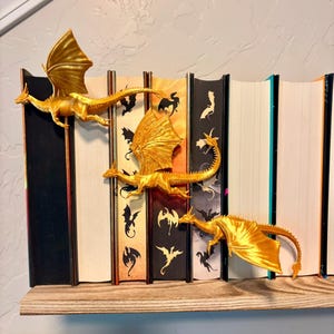 May include: Three metallic gold dragon figurines are attached to the spines of a row of books. Each dragon features spread wings, a long tail, and a slender body. The book spines display various black and white dragon silhouettes against colored backgrounds.