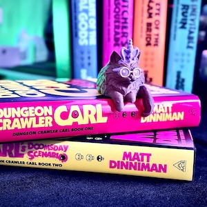 K&ouml;nnte beinhalten: Zwei B&uuml;cher mit den Titeln "Dungeon Crawler Carl" und "Carl's Doomsday Scenario" sind gestapelt. Eine Katzenfigur mit Brille und Krone sitzt auf dem oberen Buch. Die B&uuml;cher sind rosa und gelb mit schwarzem Text.