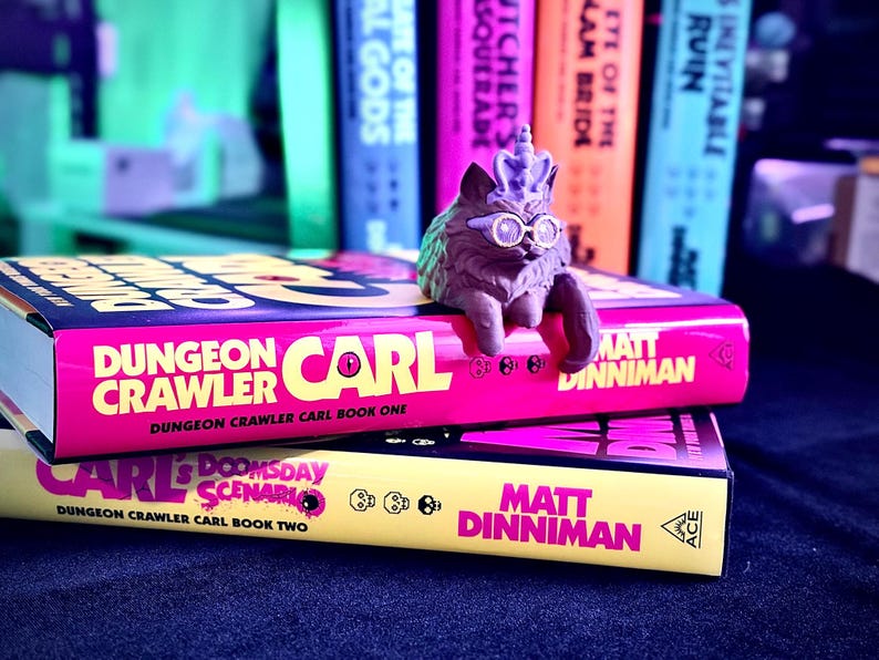 K&ouml;nnte beinhalten: Zwei B&uuml;cher mit den Titeln "Dungeon Crawler Carl" und "Carl's Doomsday Scenario" sind gestapelt. Eine Katzenfigur mit Brille und Krone sitzt auf dem oberen Buch. Die B&uuml;cher sind rosa und gelb mit schwarzem Text.
