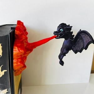 May include: A black dragon figurine with green eyes and outstretched wings, exhaling a stream of red. The dragon appears to be emerging from a fiery, orange-red structure attached to a book.