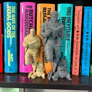 May include: Two 3D-printed figurines of a person reading a book while leaning against a creature, one in a light beige color and one in a grey color. The figurines are positioned in front of a row of colorful book spines with various titles.