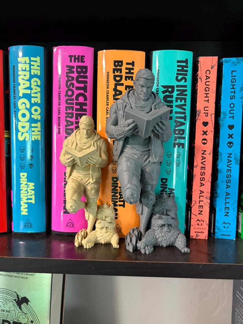 May include: Two 3D-printed figurines of a person reading a book while leaning against a creature, one in a light beige color and one in a grey color. The figurines are positioned in front of a row of colorful book spines with various titles.