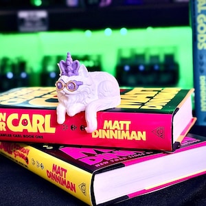 K&ouml;nnte beinhalten: Eine wei&szlig;e Katzenfigur mit lila Akzenten, die eine Sonnenbrille und eine Krone tr&auml;gt, liegt auf zwei B&uuml;chern. Die B&uuml;cher haben pinkfarbene und gelbe Einb&auml;nde mit dem Namen des Autors, Matt Dinniman, und dem Titel "Dungeon Crawler Carl".