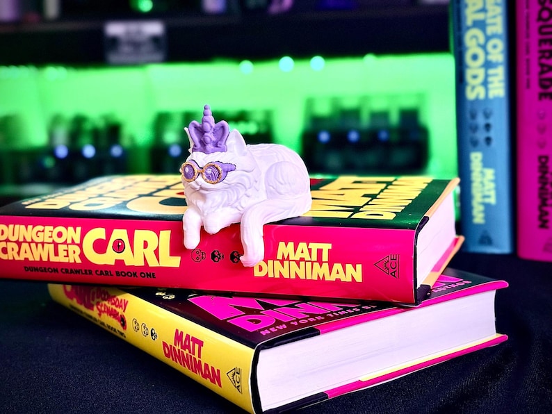 K&ouml;nnte beinhalten: Eine wei&szlig;e Katzenfigur mit lila Akzenten, die eine Sonnenbrille und eine Krone tr&auml;gt, liegt auf zwei B&uuml;chern. Die B&uuml;cher haben pinkfarbene und gelbe Einb&auml;nde mit dem Namen des Autors, Matt Dinniman, und dem Titel "Dungeon Crawler Carl".