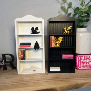 May include: Two miniature bookshelves, one white and one black, are displayed on a wooden surface. The white shelf holds books and dragon figurines. The black shelf also has books and a golden dragon figurine. A decorative sign reads "to be read."
