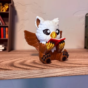 May include: A figurine of a seated owl-like creature with white head and chest, brown wings and body, and black feet. It holds a small red book with gold pages. The figurine is on a wooden surface.