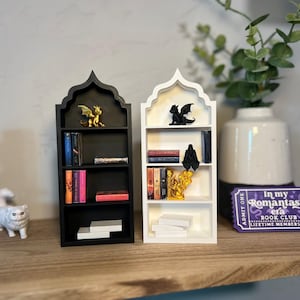 May include: Miniature black and white bookshelves with arched tops, each holding books and decorative figurines of dragons. A purple ticket with text "In my Romantasy era Book Club" sits on the shelf. A white cat figurine and a small rolling bookshelf are also present.