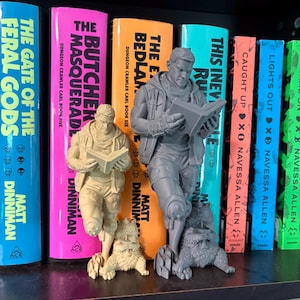May include: Two 3D printed figurines of a character reading a book while standing next to a small creature. One figurine is light beige and the other is grey. Both figures feature detailed clothing, including a vest and scarf, and are posed standing.