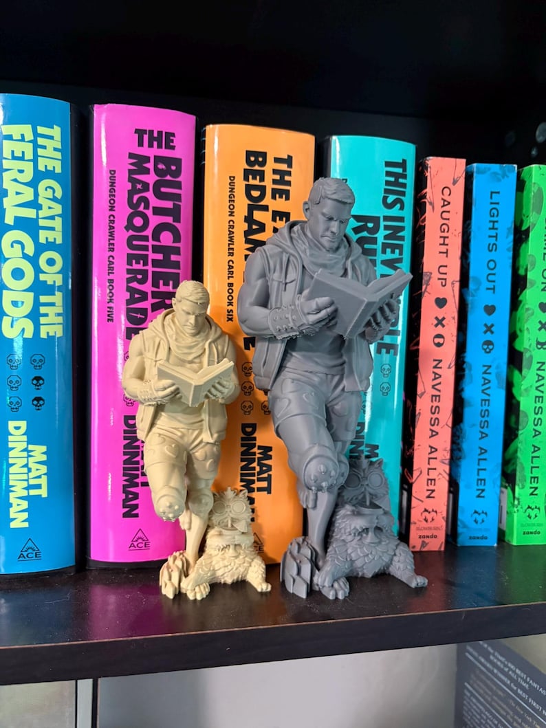 May include: Two 3D printed figurines of a character reading a book while standing next to a small creature. One figurine is light beige and the other is grey. Both figures feature detailed clothing, including a vest and scarf, and are posed standing.