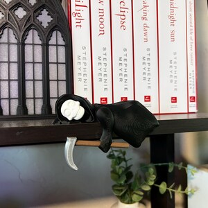 May include: A black and white Grim Reaper figurine with a scythe, resting on a dark shelf. The figurine has a white skull head and is draped in a black cloak. Books with red and white spines are in the background.