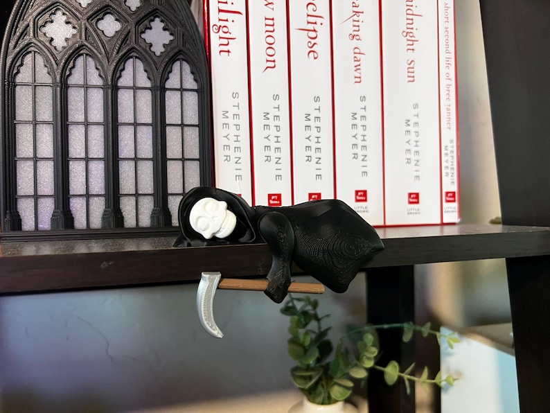 May include: A black and white Grim Reaper figurine with a scythe, resting on a dark shelf. The figurine has a white skull head and is draped in a black cloak. Books with red and white spines are in the background.