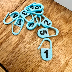 May include: A collection of light blue stitch markers with black numbers 1 through 5. The stitch markers are in the shape of a safety pin. The markers are scattered on a wooden surface.