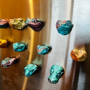 Pode incluir: Uma coleção de 11 ímãs de geladeira de cabeça de réptil impressos em 3D e coloridos. Os ímãs são em vários tons de verde, azul, amarelo e laranja.
