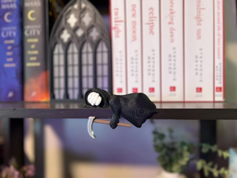 May include: A black Grim Reaper figurine with a white face and a scythe. The figurine is resting on a dark shelf, with books and a decorative gothic window in the background. The figurine is a unique decorative item.