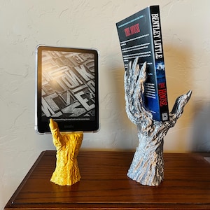May include: Two 3D printed bookends in the shape of a clawed hand, one gold and one silver, holding a book and an e-reader. The book is titled "The House" by Bentley Little.