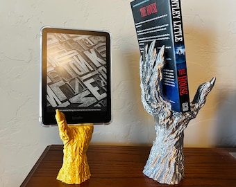 May include: Two 3D printed bookends in the shape of a clawed hand, one gold and one silver, holding a book and an e-reader. The book is titled "The House" by Bentley Little.